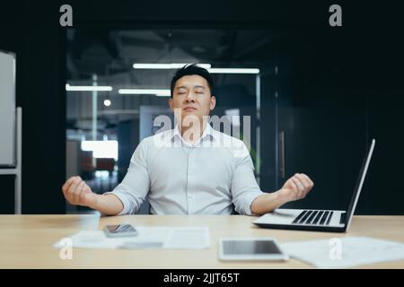 Una pausa al lavoro. Bel giovane uomo d'affari asiatico meditando sul posto di lavoro in ufficio, chiuso gli occhi, tiene le mani in posizione lotus, si riposa, si rilassa. Foto Stock