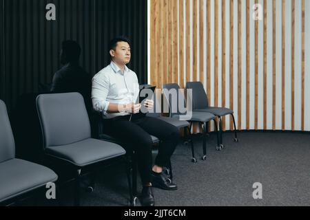 Un giovane e bell'uomo asiatico sta aspettando nella sala di ricevimento per un'intervista per un lavoro in un ufficio, banca, azienda d'affari. Tiene una cartella nelle sue mani, si preoccupa, la scuote Foto Stock