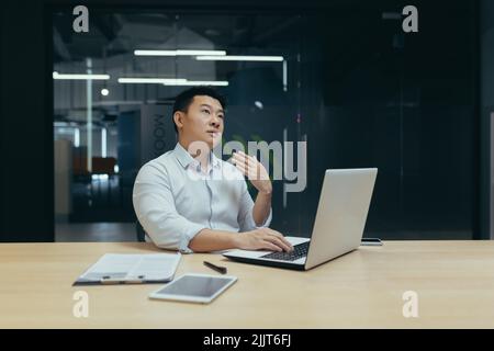 Giovane uomo d'affari asiatico che non si sente bene al lavoro. Lui ondeggia la mano, fa caldo, ha poco respiro. Seduti alla scrivania in ufficio Foto Stock