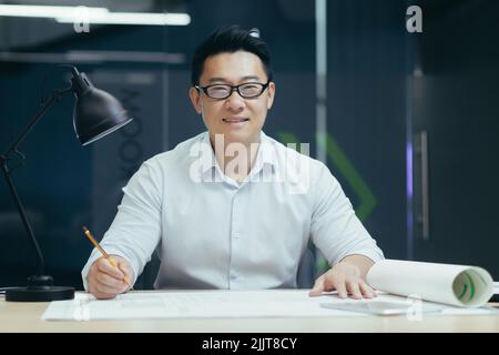 Ritratto del giovane uomo asiatico indossare occhiali, designer, architetto, guardando la macchina fotografica, sorridente. Seduti a un tavolo in un ufficio moderno, lavorando con i disegni, tenendo una matita nelle mani. Foto Stock