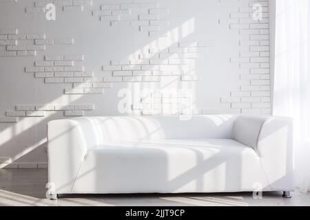 Interno luminoso e minimalista con divano bianco accanto alla finestra e sgabello nero Foto Stock