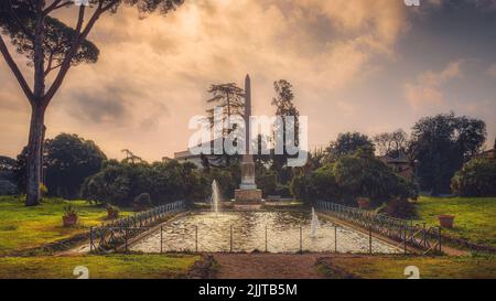 Una vista panoramica delle fontane e un obelisco in un giardino verde di Villa Torlonia a Roma, Italia al tramonto Foto Stock