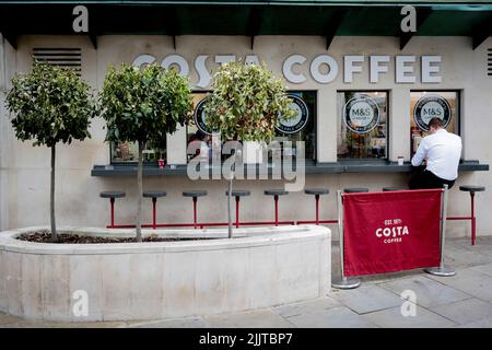 Accanto a tre arbusti che si appassiscono per la mancanza di acqua, un uomo siede su uno sgabello in un bar da asporto Costa Coffee nella City di Londra, il quartiere finanziario della capitale, il 27th luglio 2022, a Londra, Inghilterra. Costa Coffee è stata fondata a Londra nel 1971 da Sergio Costa - acquisita da Whitbread nel 1995 poi venduta nel 2019 alla Coca-Cola Company in un accordo del valore di £ 3,9bn. La sua attività nel Regno Unito ha 2.121 ristoranti nel Regno Unito, oltre 6.000 distributori automatici Costa Express e altri 1.280 punti vendita all'estero. Foto Stock