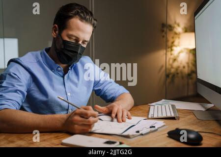 Giovane uomo bianco in maschera protettiva che scrive le note mentre lavora in ufficio Foto Stock