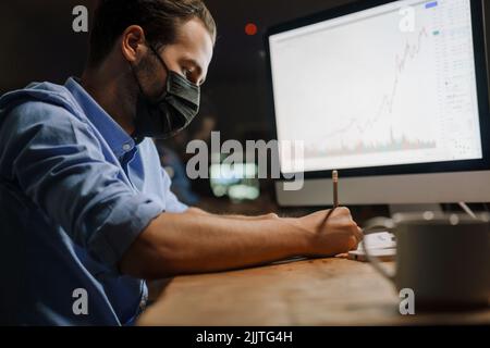 Giovane uomo bianco in maschera protettiva che scrive le note mentre lavora in ufficio Foto Stock