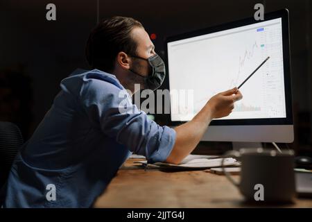 Giovane uomo bianco in maschera protettiva che lavora con il computer in ufficio Foto Stock