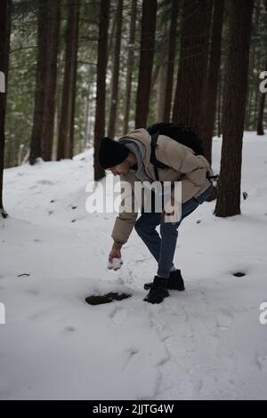 Un colpo verticale di un giovane caucasico che fa una palla di neve in una foresta invernale Foto Stock
