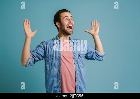 Giovane uomo eccitato che indossa una camicia urlando con le mani isolate su sfondo blu Foto Stock