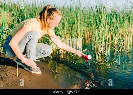 Giovane volontario femminile soddisfatto di raccogliere rifiuti, una bottiglia di plastica e tazze di caffè, pulire la spiaggia con un lago. Donna raccolta rifiuti in borsa Foto Stock