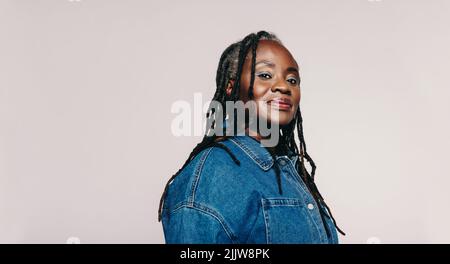 Donna alla moda con dreadlock che guarda la fotocamera in piedi su uno sfondo grigio. Ritratto di una donna matura che indossa una giacca in denim e un Foto Stock