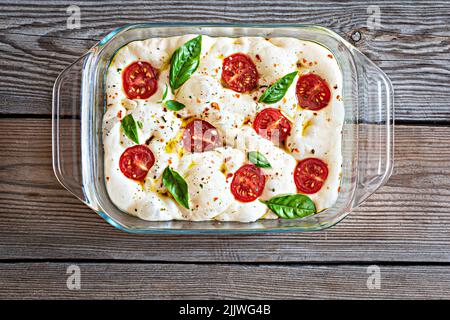 Pane fresco piatto italiano (crudo) Focaccia con pomodori, olive, aglio ed erbe su sfondo di legno. Foto Stock