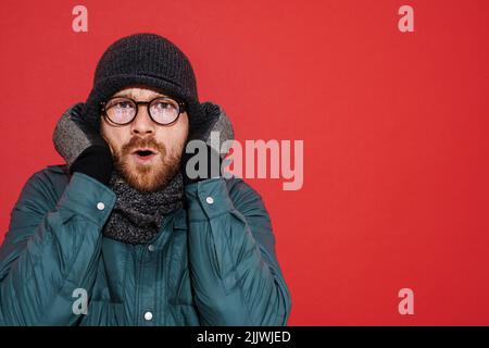 Uomo zenzero che indossa cappello e giacca tremante e sensazione di freddo isolato su sfondo rosso Foto Stock