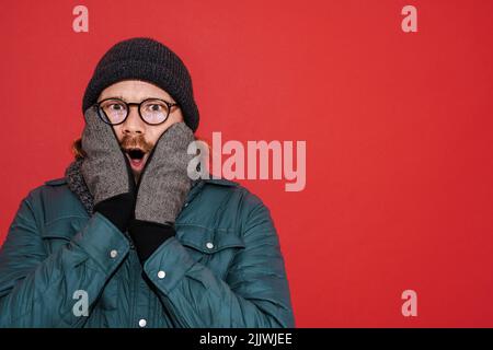 Uomo zenzero che indossa mittens e giacca che esprime sorpresa isolato su sfondo rosso Foto Stock