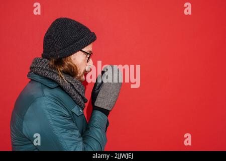 Uomo zenzero che indossa cappello e giacca tremante e sensazione di freddo isolato su sfondo rosso Foto Stock