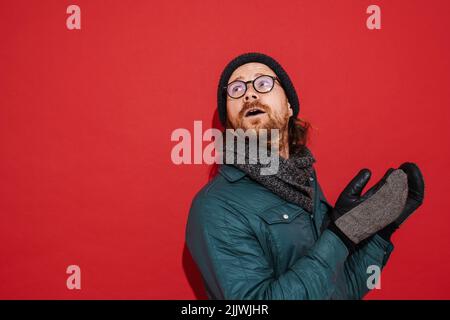 Uomo zenzero che indossa cappello e giacca tremante e sensazione di freddo isolato su sfondo rosso Foto Stock