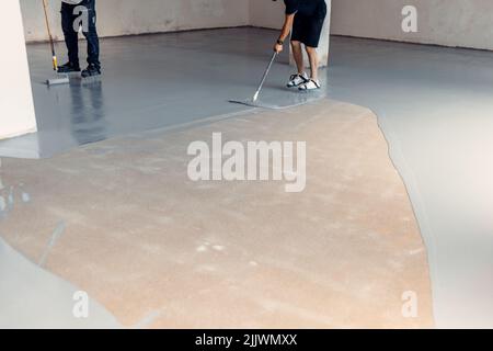 Due operai edili che applicano resina epossidica grigia in un ambiente industriale Foto Stock