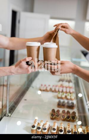 Vista ritagliata del venditore che dà la borsa di carta e tazze al cliente africano americano vicino vetrina in pasticceria Foto Stock