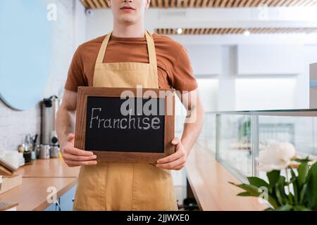 Vista croppata del venditore che tiene la lavagna con la scritta in franchising vicino alla vetrina e fiore nel negozio di dolci Foto Stock