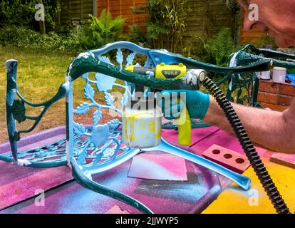 Uomo che usa pistola a spruzzo di vernice per spruzzare e upcycle sedie da giardino in ferro battuto, UK Foto Stock