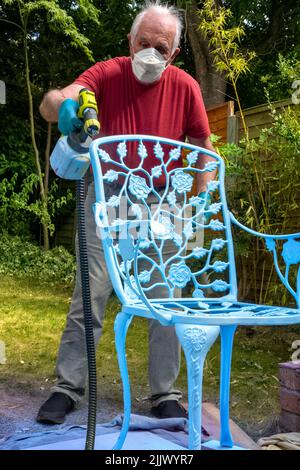 Uomo anziano che usa la pistola a spruzzo per spruzzare e rifare sedie da giardino in ferro battuto, Regno Unito Foto Stock
