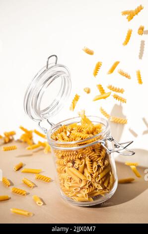 Alto angolo di mazzetto di fusilli non cotti e penne di pasta che cadono in vaso di vetro aperto inclinando sul tavolo su sfondo bianco Foto Stock