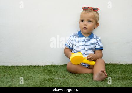 il ragazzino di 1 anni siede su un'erba verde con occhiali da sole. Foto Stock