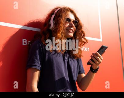 Giovane donna sorridente con cuffie che scorrono attraverso lo smartphone contro la parete arancione Foto Stock