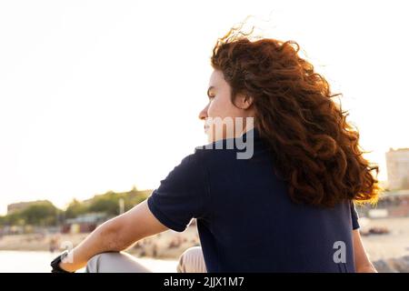 Vista posteriore della giovane donna con testa rossa seduta contro la spiaggia in città Foto Stock