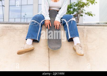 Donna ritagliata e irriconoscibile in abbigliamento casual con skateboard guardando lontano mentre si siede sul bordo di cemento con skate Park in città Foto Stock