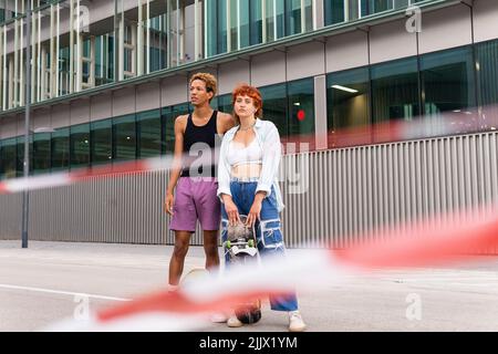 Coppia di amici multirazziali con skateboard in piedi sul marciapiede vicino edificio sulla strada con blurred e nastro di protezione bianco in città Foto Stock