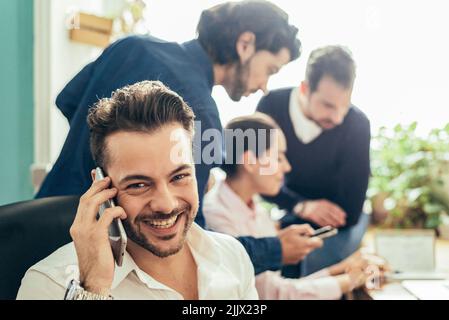 buon lavoratore di sesso maschile in abbigliamento formale che ha una conversazione telefonica vicino a un gruppo di colleghi mentre lavora insieme in un moderno ufficio leggero Foto Stock