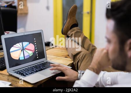 Vista laterale di un lavoratore di sesso maschile concentrato che naviga su netbook con grafici mentre lavora su un progetto aziendale in un ambiente di lavoro moderno e leggero Foto Stock