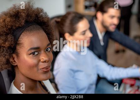 Una donna afro-americana positiva che guarda la macchina fotografica mentre si siede vicino a un gruppo di colleghi in uno sfondo sfocato in un ufficio leggero Foto Stock