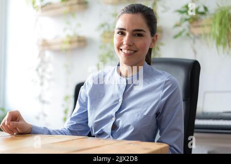 Allegra manager caucasica femminile in blusa bianca con capelli ricci sorridenti e guardando la macchina fotografica mentre si siede dietro il tavolo in ufficio illuminato dal sole Foto Stock