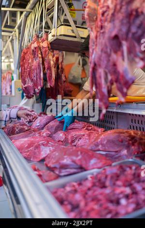 Anonimo uomo ispanico in uniforme e guanti di lattice prendendo pezzo di carne cruda dalla stalla mentre servendo cliente femminile di mezza età che punta via nel butche Foto Stock