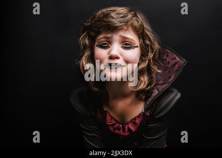 Ragazza piccola sconvolta con capelli ricci e trucco scuro in costume incantatrice che piange e guardando la fotocamera su sfondo nero durante Halloween holida Foto Stock