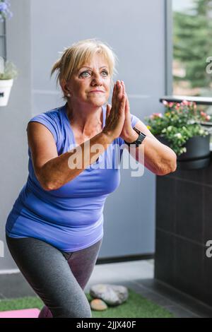 Donna anziana focalizzata pratica yoga con le mani afferrate guardando lontano in balcone Foto Stock