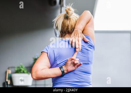 Vista posteriore della donna anziana che pratica yoga in posizione fronte mucca o gomukhasana durante l'allenamento a casa Foto Stock