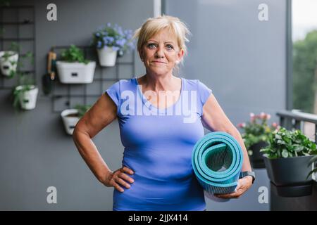 Ritratto di donna anziana sorridente con tappeto di yoga arrotolato in palestra Foto Stock