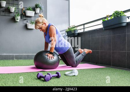 Vista laterale completa del corpo della donna anziana che fa l'allenamento con la palla fitness sul balcone Foto Stock