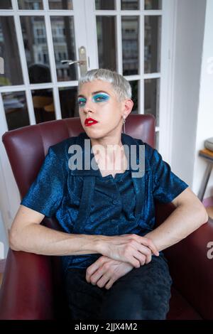 Ritratto di uomo androgino sicuro che indossa il trucco seduta su poltrona a casa Foto Stock