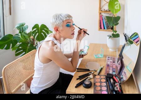 Uomo transgender che indossa un ombretto blu che applica mascara mentre guarda nello specchio Foto Stock