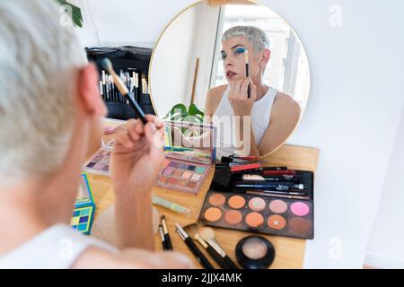 Uomo transgender che indossa un ombretto blu che applica mascara mentre guarda nello specchio Foto Stock