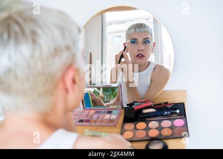 Uomo transgender che indossa un ombretto blu che applica mascara mentre guarda nello specchio Foto Stock