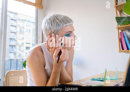 Uomo transgender che indossa un ombretto blu che applica mascara mentre guarda nello specchio Foto Stock