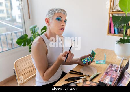 Uomo transgender che indossa un ombretto blu che applica mascara mentre guarda la macchina fotografica Foto Stock