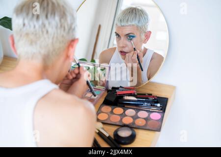 Uomo transgender che indossa un ombretto blu che applica mascara mentre guarda nello specchio Foto Stock