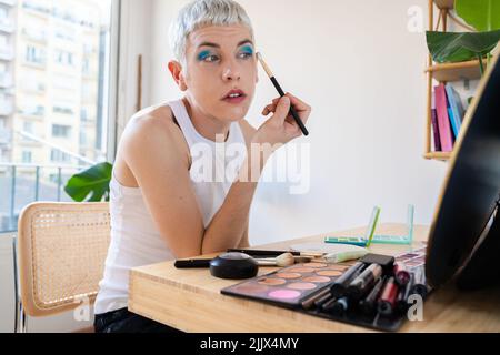 Uomo transgender che indossa un ombretto blu che applica mascara mentre guarda nello specchio Foto Stock