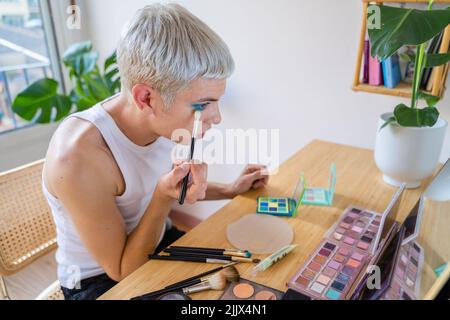 Uomo transgender che indossa un ombretto blu che applica mascara mentre guarda nello specchio Foto Stock