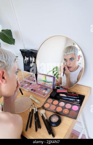 Uomo transgender che indossa un ombretto blu che applica mascara mentre guarda nello specchio Foto Stock